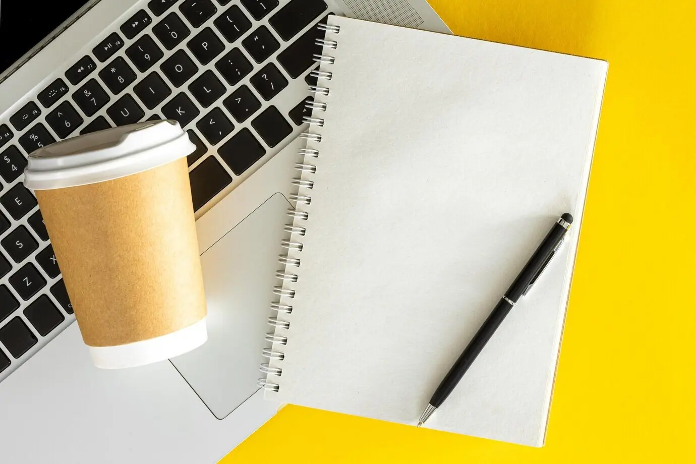 Unbeschriebener Notizblock, Laptop und Tasse Kaffee auf gelbem Hintergrund als Flatlay.